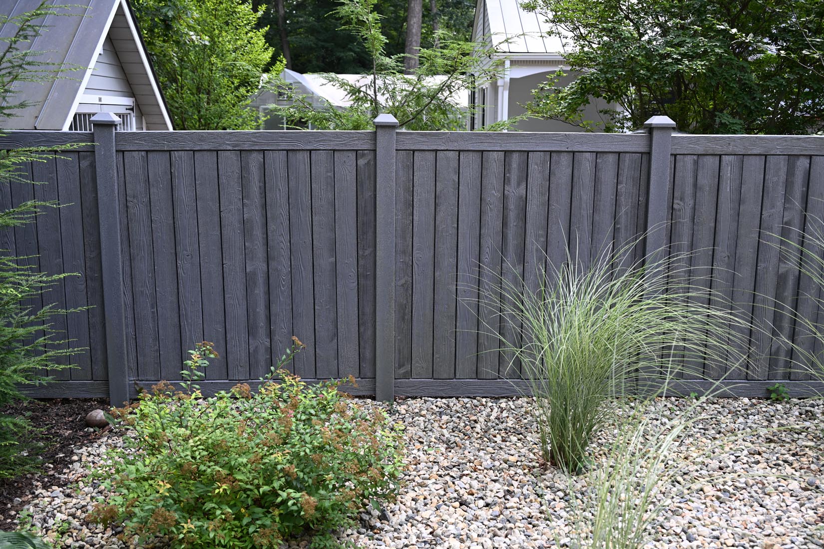 A tall, gray wooden fence stands behind a garden bed with decorative rocks, a green shrub, and ornamental grasses. Trees and houses are visible in the background.
