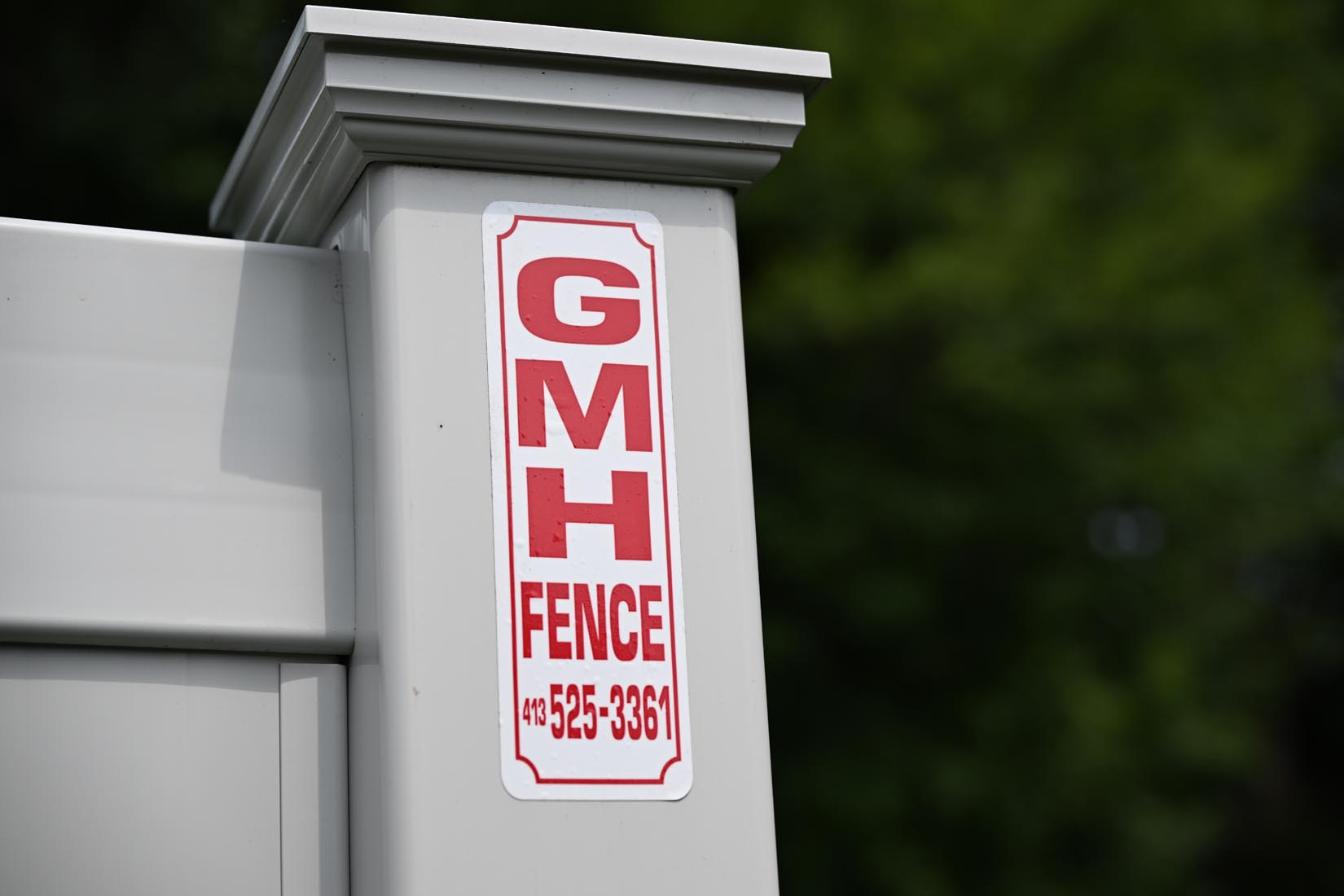 GMH Fence Residential Fencing Western MA 187 A close-up of a white fence post with a sign reading GMH FENCE and a phone number in red letters, set against a blurred green background.