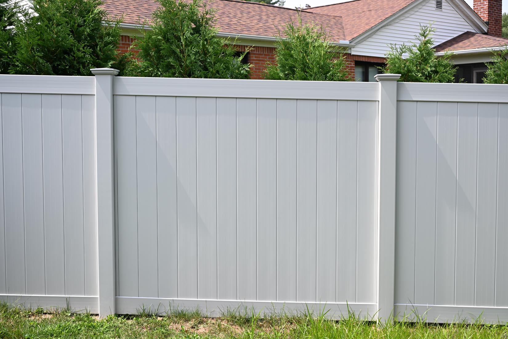 A white vinyl privacy fence at Enfield, CT home stands in front of green shrubs, with part of a brick house and a shingled roof visible in the background.