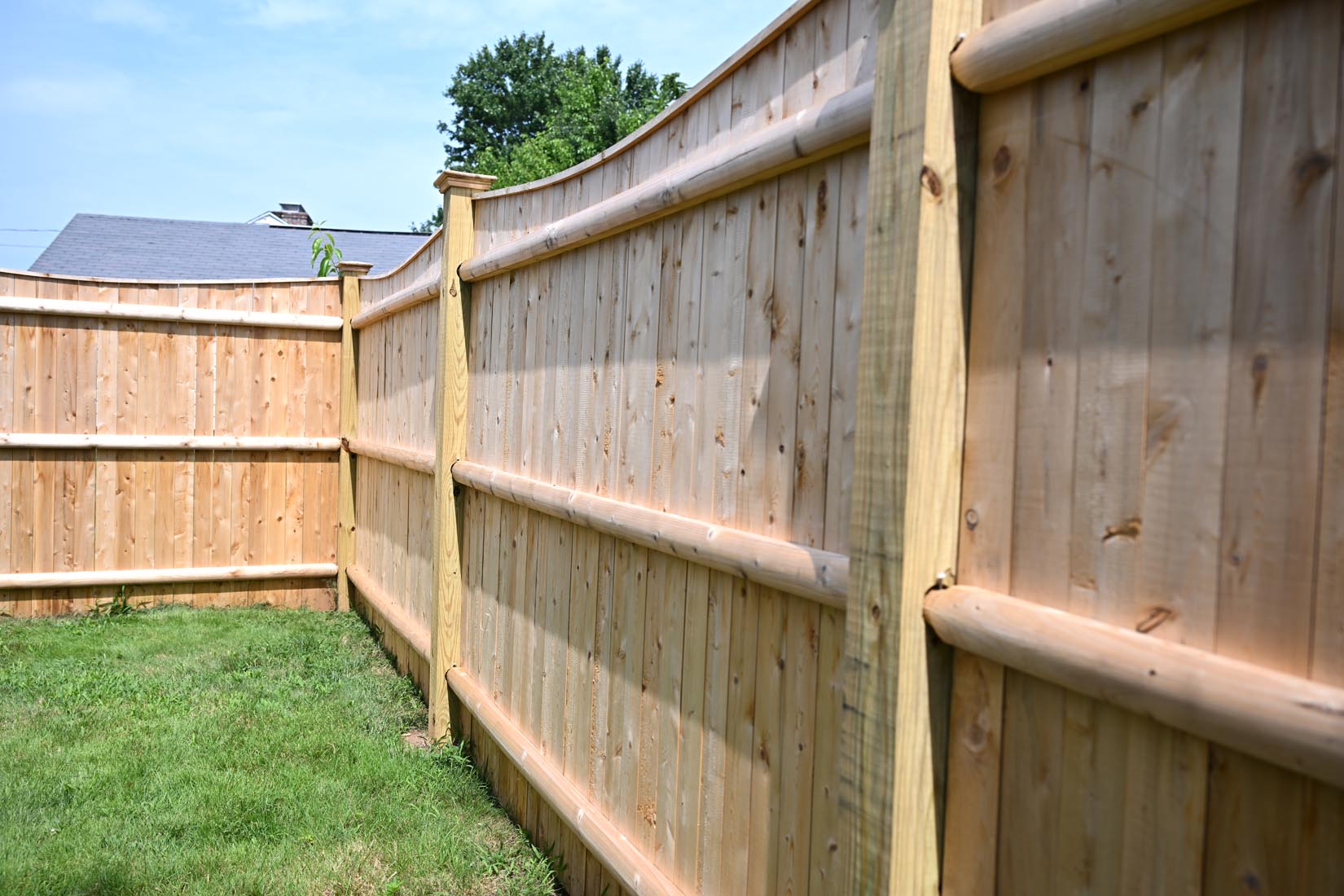A close-up view of a wooden privacy fence with vertical planks and horizontal supports, enclosing a grassy backyard under a partly cloudy sky. The fence curves slightly at the top.