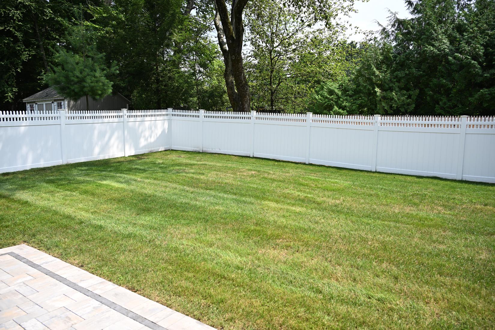 GMH Fence Residential Fencing Western MA 241 A backyard with a neatly mowed green lawn, bordered by a white picket fence, with trees and a shed in the background and a paved patio in the foreground.