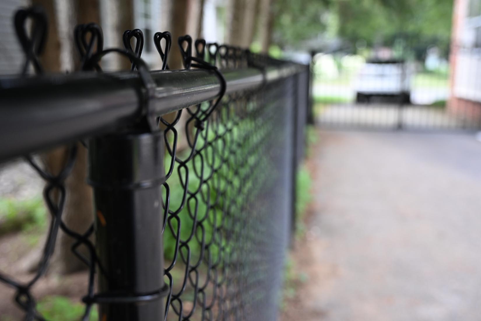 Close-up of a black chain-link fence along a paved path, with trees and a blurred gate in the background.