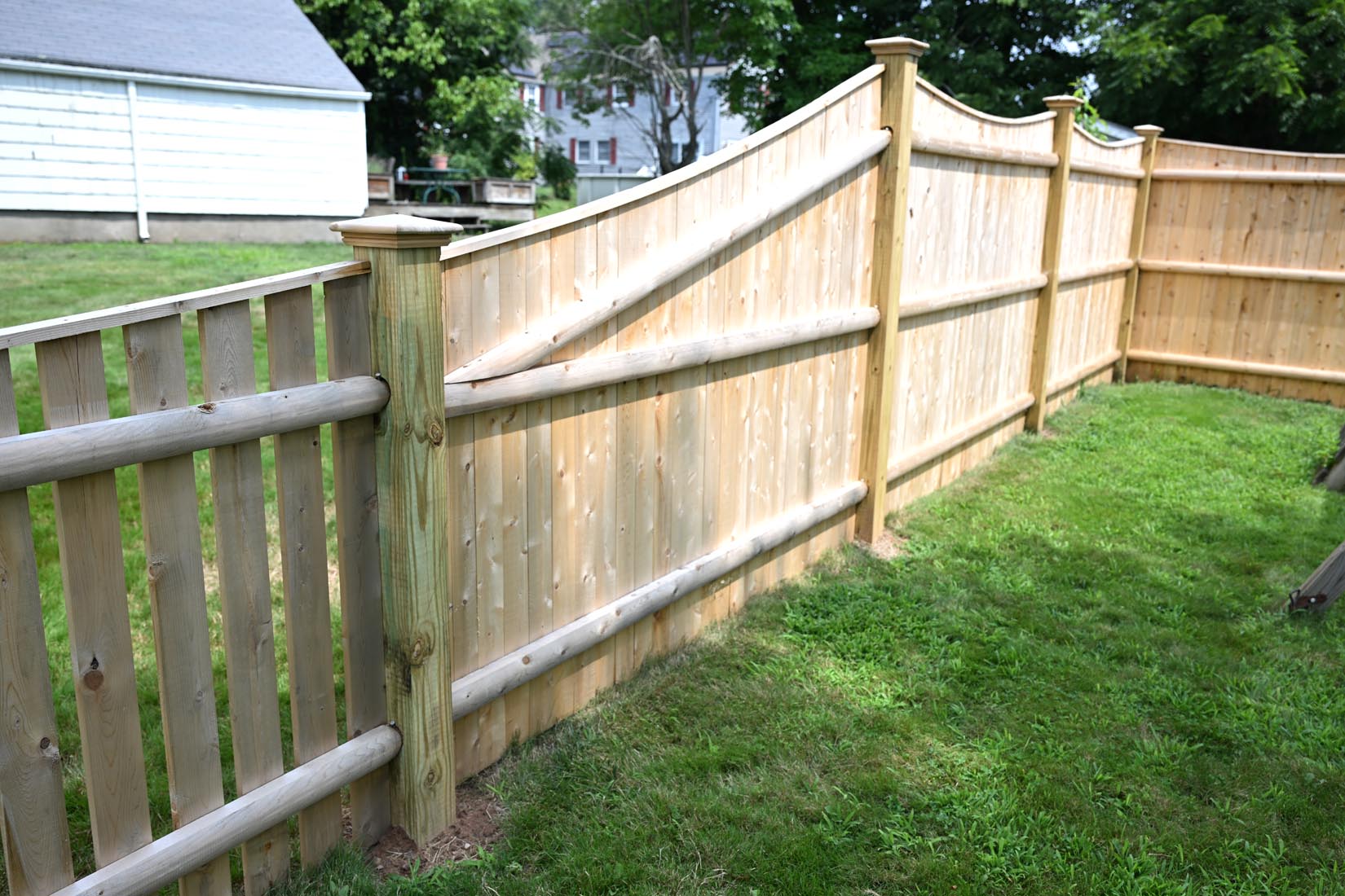 A new wooden privacy fence with a decorative top edge runs along a green, grassy backyard, separating it from neighboring properties. Trees and houses are visible in the background.