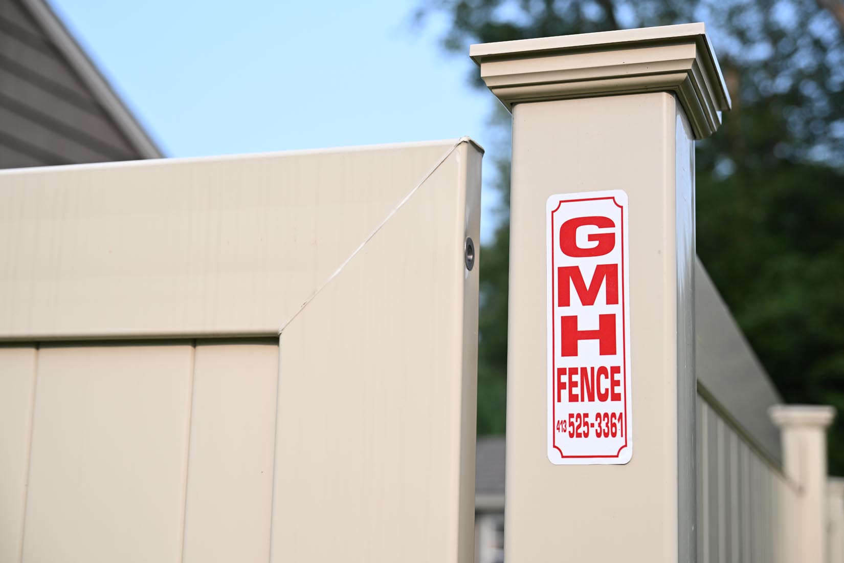A beige vinyl fence with a red and white GMH Fence sign and phone number attached to a post; the background includes a blurry house and green trees. Taken at Enfield, CT home.