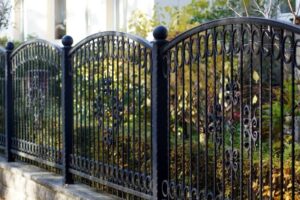 Black ornamental and decorative fencing--aluminum or steel material with swirls and finials stands in front of a lush garden, sunlight shining on the foliage and fence