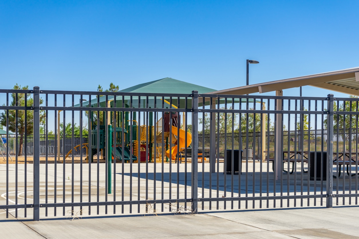 A closed school playground A playground with a yellow slide and green canopy is seen behind a black metal fence on a sunny day. Picnic tables are under a covered area nearby. The ground is paved and trees are in the background.