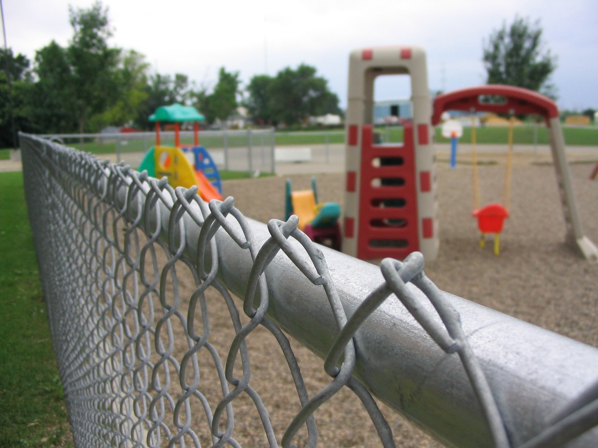 Grass next to children's playground A close-up of a chain-link fence with a playground in the background. The playground features colorful slides, climbing structures, and swings, all set on a gravel surface with trees and grass nearby.