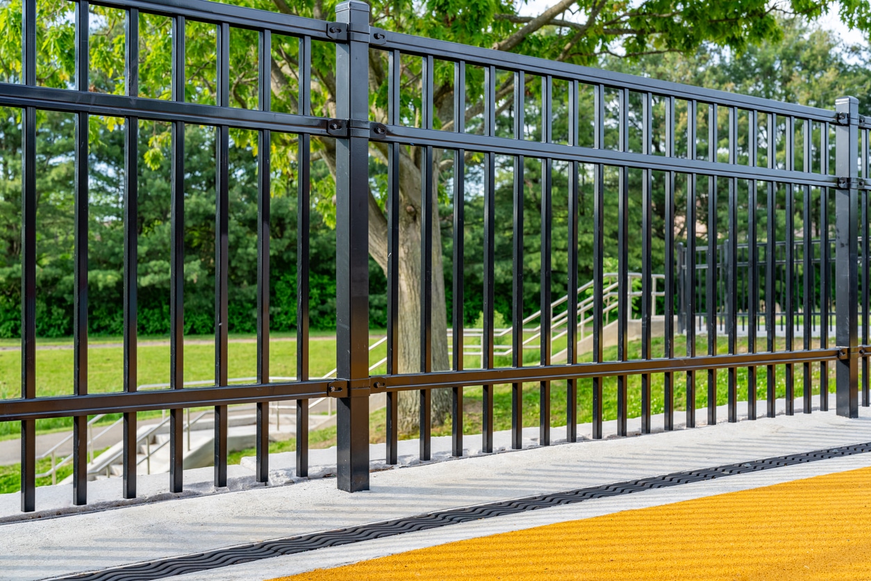 Late afternoon image of an ornamental metal trench drain in concrete with shadow pattern from an adjacent ornamental fence. Black metal fence with vertical bars on a concrete base, yellow textured strip in the foreground, and a green park with trees and shrubs in the background.