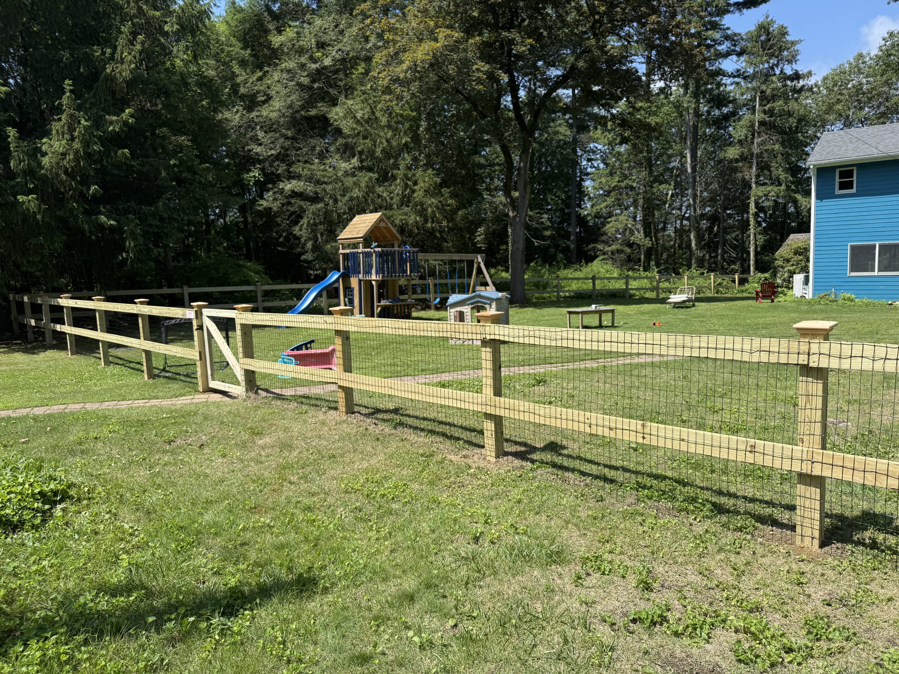 Wood and wire fence at family home in Wilbrham, MA. Fence features brown cedar wood, dark mesh wire. Playset with green slide and swings in background with small view of blue siding on house.