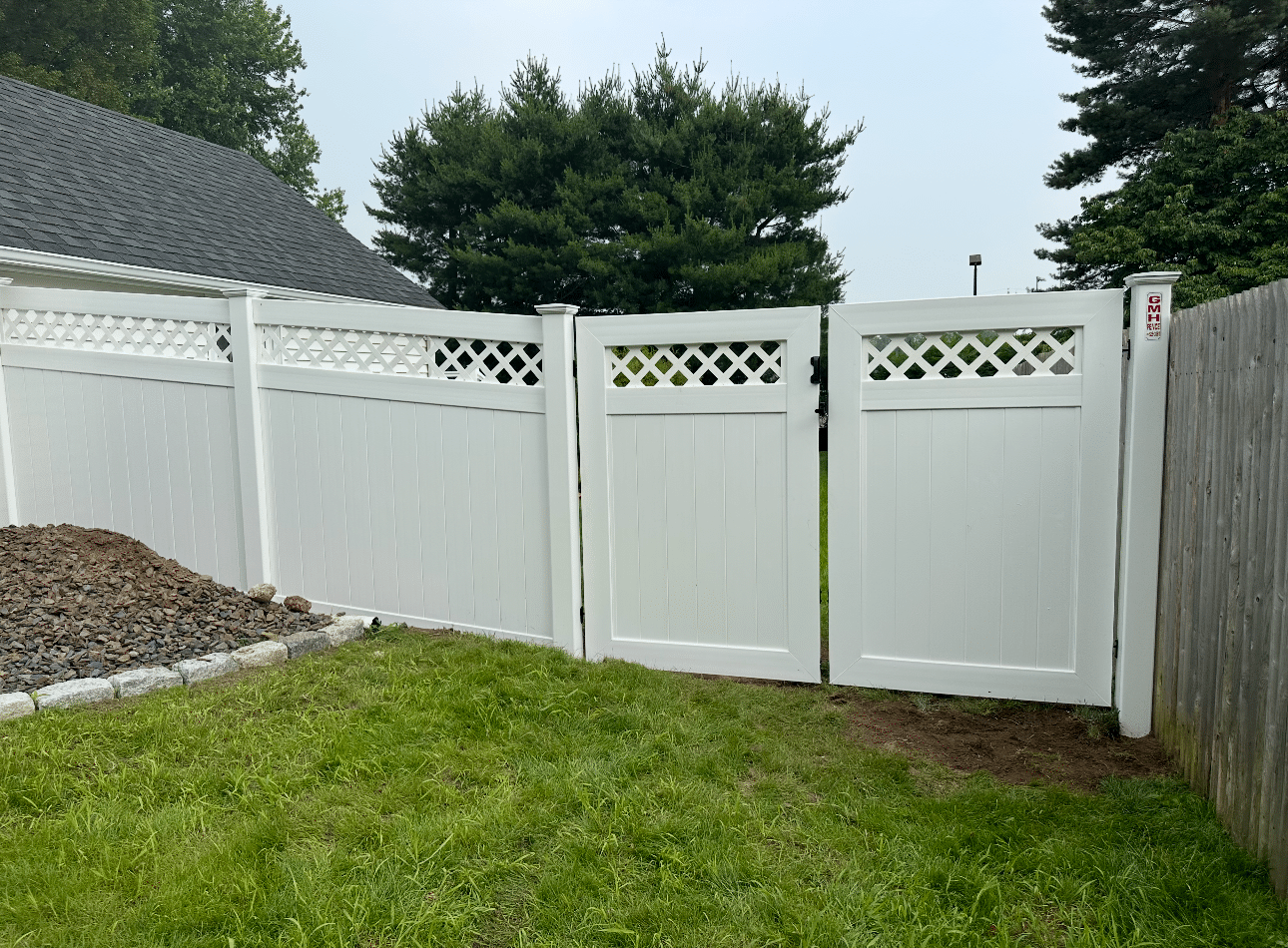 White vinyl fence with two gates and decorative lattice tops stands between a gray-sided building and a wooden fence, enclosing a grassy yard bordered by a small pile of rocks and trees in the background.