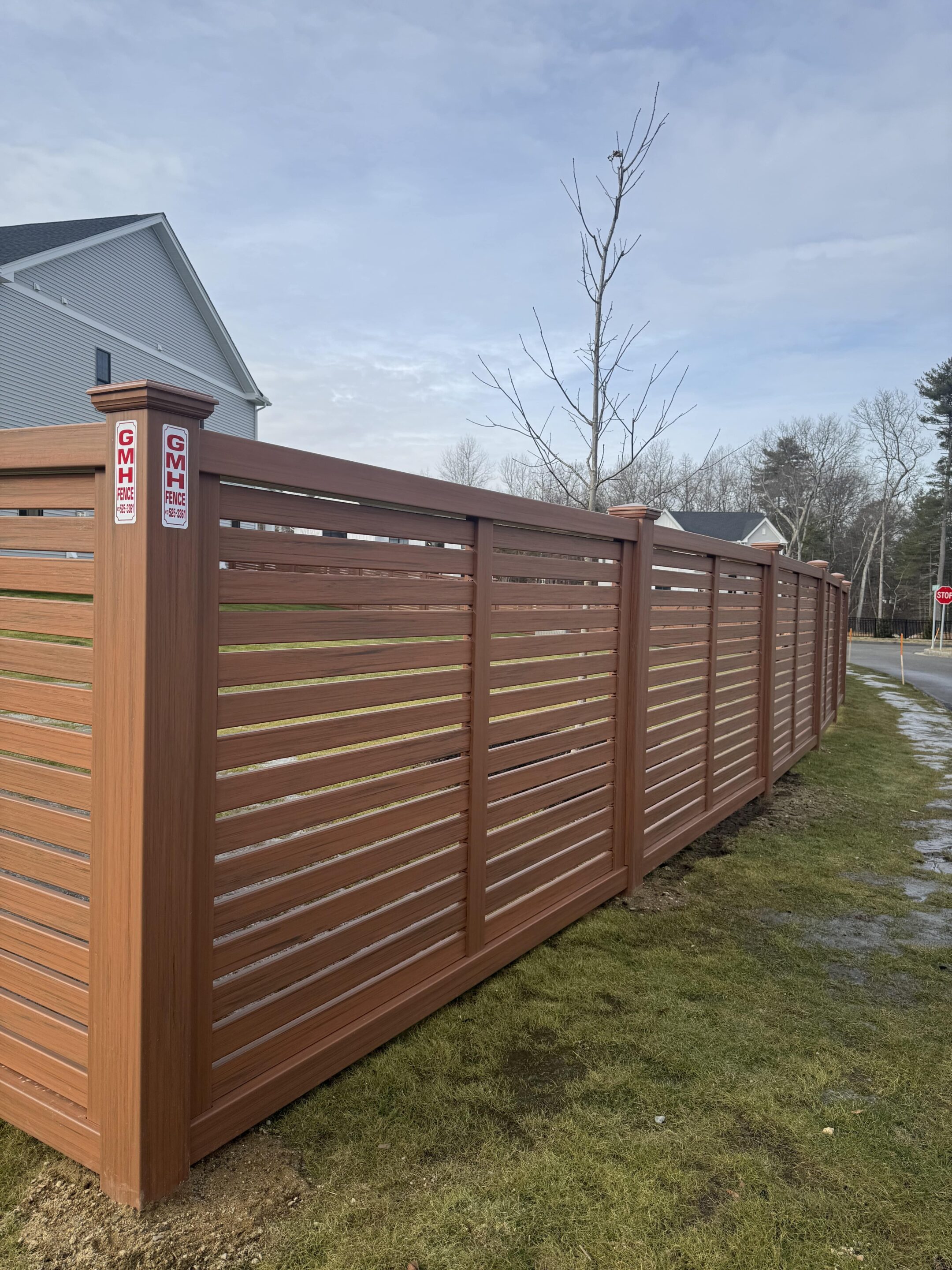 A tall wood-like vinyl fence with horizontal slats stands beside a grassy yard; a leafless tree grows near the fence, and houses are visible in the background under a cloudy sky. Captured in Hudson, MA.
