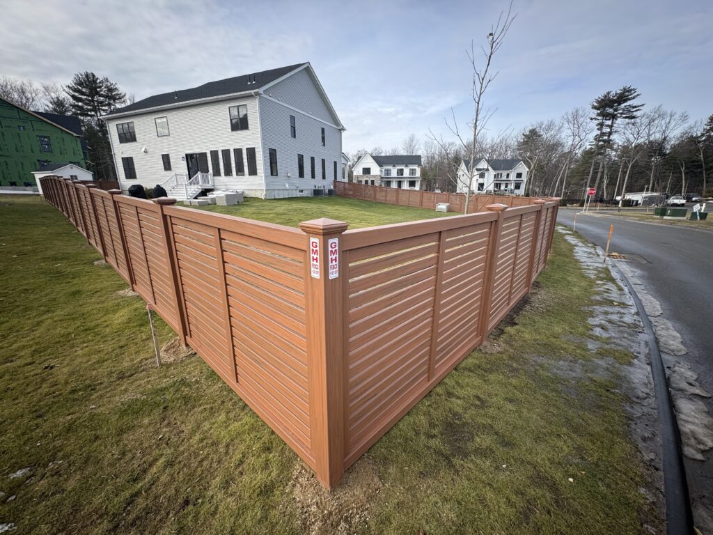 A brown wooden privacy fence forms a corner around the yard of a white house, with a street and other houses visible in the background under a partly cloudy sky.