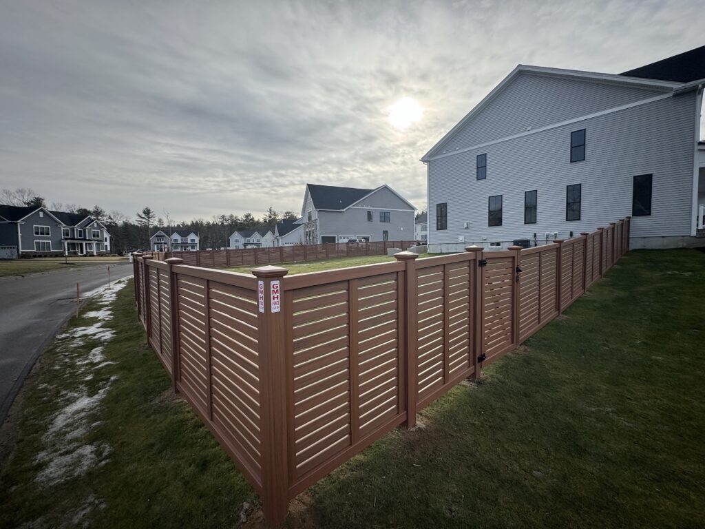 A brown slatted vinyl fence encloses a grassy yard next to a modern gray house in a suburban neighborhood under a cloudy sky with the sun partially visible in Hudson, MA. Other houses and a street are in the background.