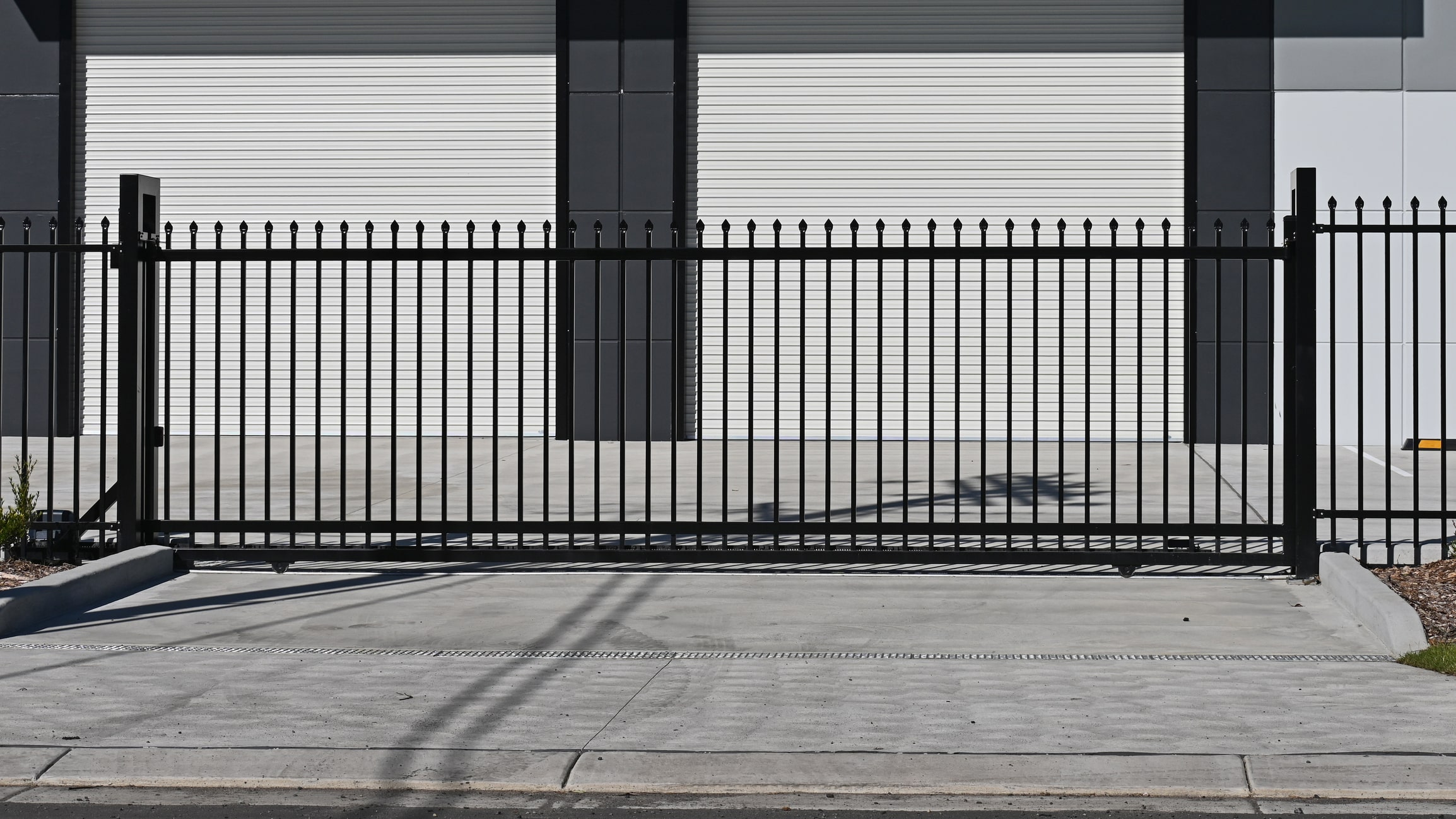 A black metal fence with vertical bars stands in front of a building with three large, closed white roller doors. The concrete sidewalk and driveway are visible in the foreground.