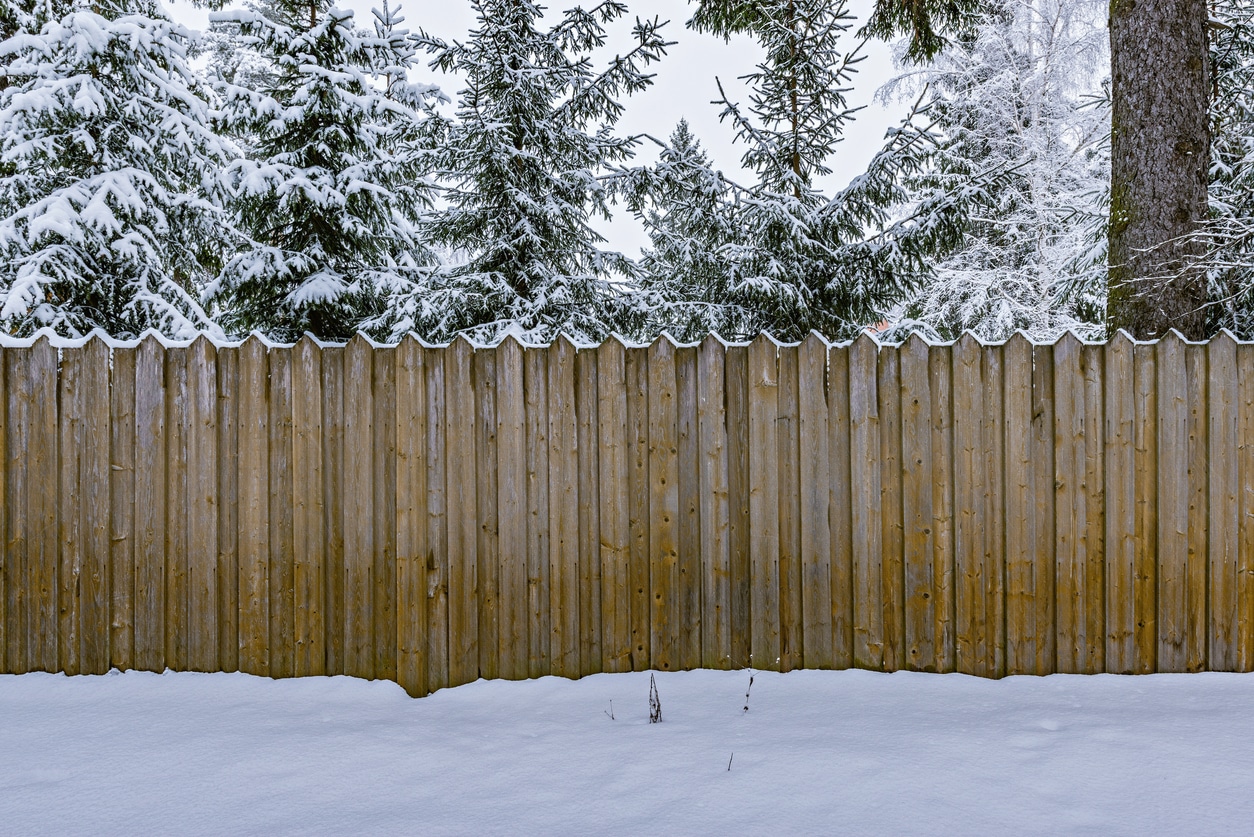 On a winter day in MA & CT, a wooden fence stands quietly before snow-covered evergreen trees, with fresh snow blanketing the ground and the top of the fence, illustrating wood fence repair.