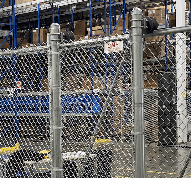A chain-link fence with a GMH sign stands in front of tall metal warehouse shelves filled with cardboard boxes. Taken in Enfield, CT.