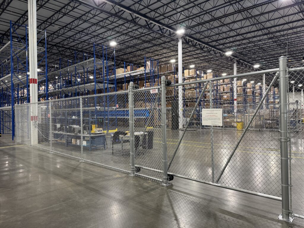 Large warehouse interior with tall blue metal shelving, stacks of boxes, and a chain-link fence with a locked gate in the foreground. The concrete floor and ceiling with lights are also visible.