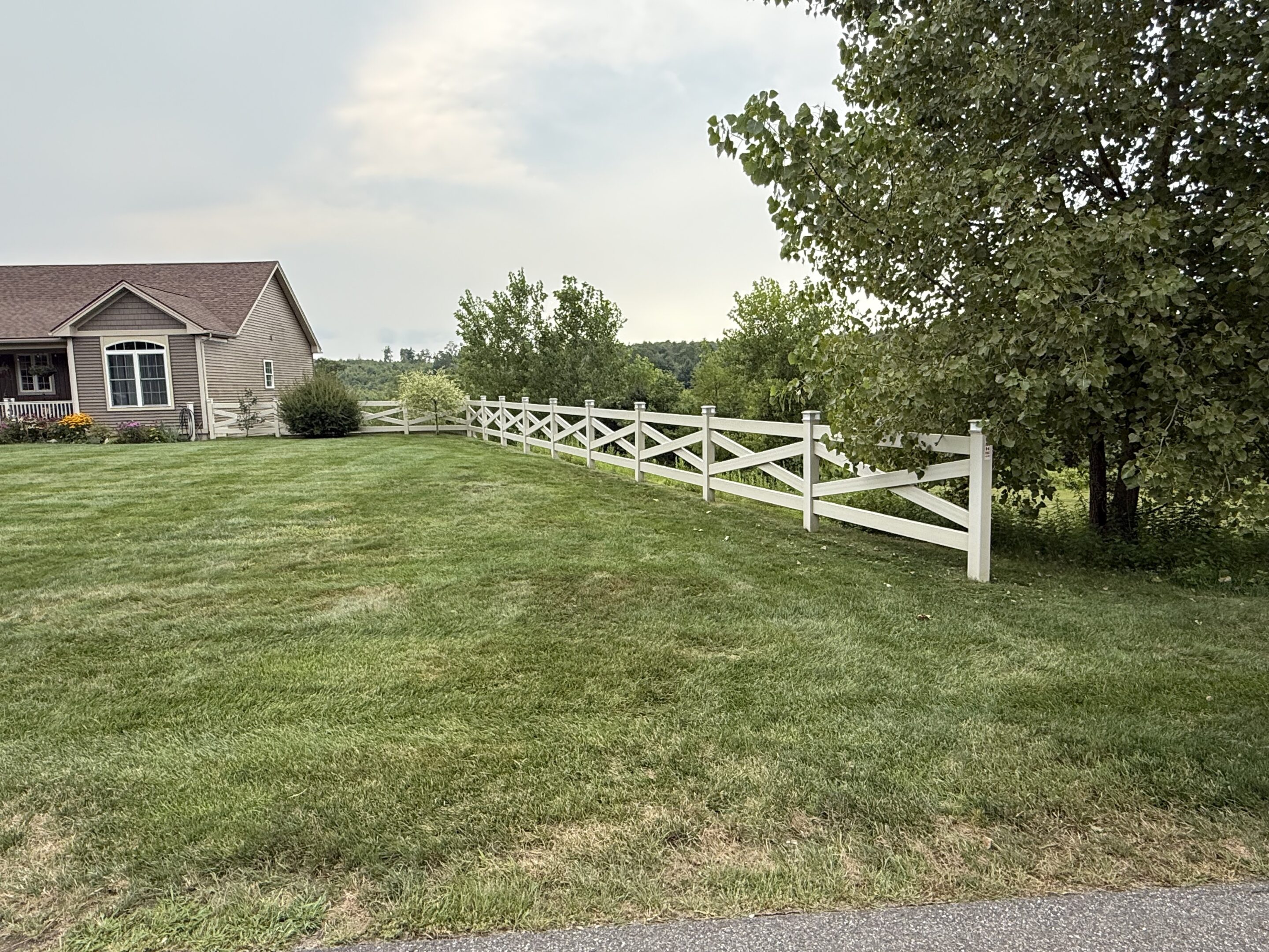 A white wooden fence runs along the edge of a neatly mowed lawn beside a house with tan siding. Trees border the fence, and the sky is partly cloudy.