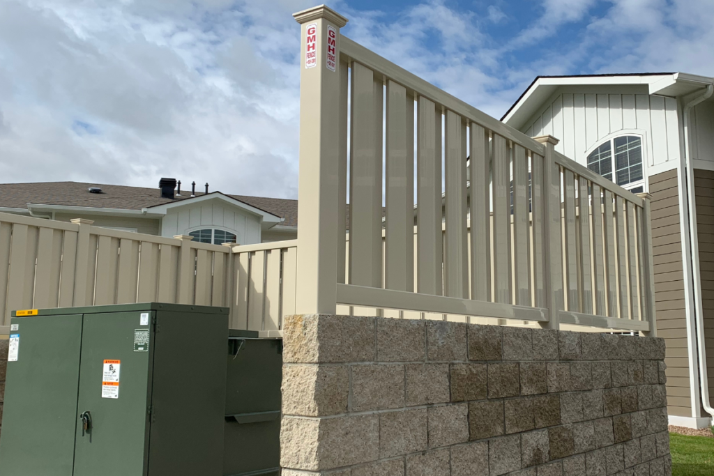 A beige fence sits atop a short stone wall beside a green utility box, with a modern house featuring large windows and tan siding in the background under a partly cloudy sky.
