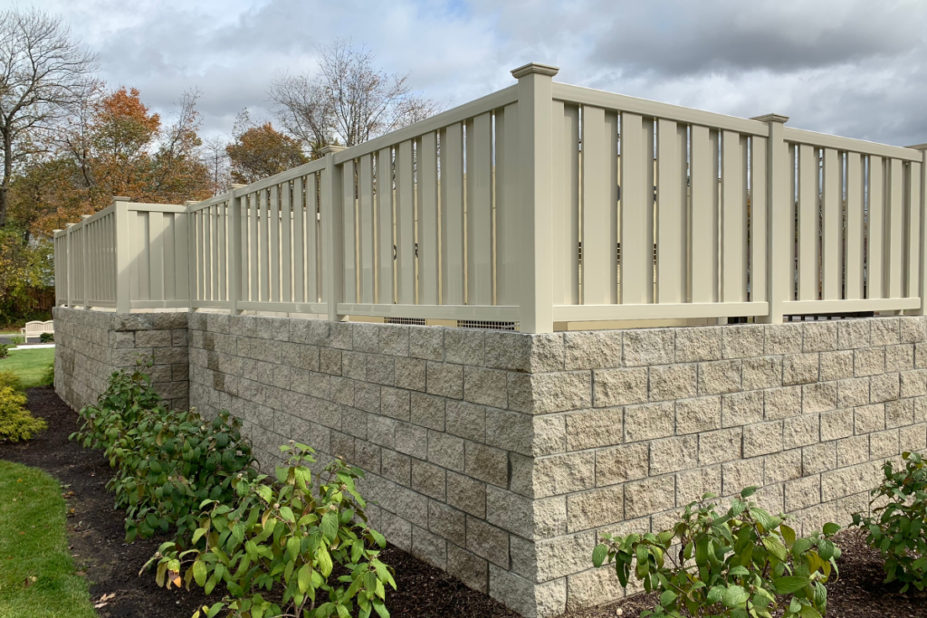 A beige vinyl fence sits atop a retaining wall made of large, stacked concrete blocks. Green shrubs and mulch line the base, with trees and a cloudy sky in the background.