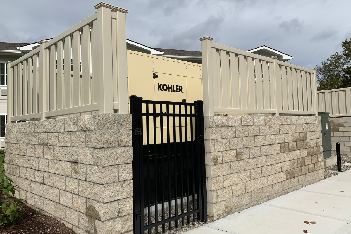 A beige Kohler generator in Waterbury, CT is enclosed by light tan fencing and stone walls with a black metal gate, situated on a sidewalk near a building with gray and white siding under a cloudy sky.