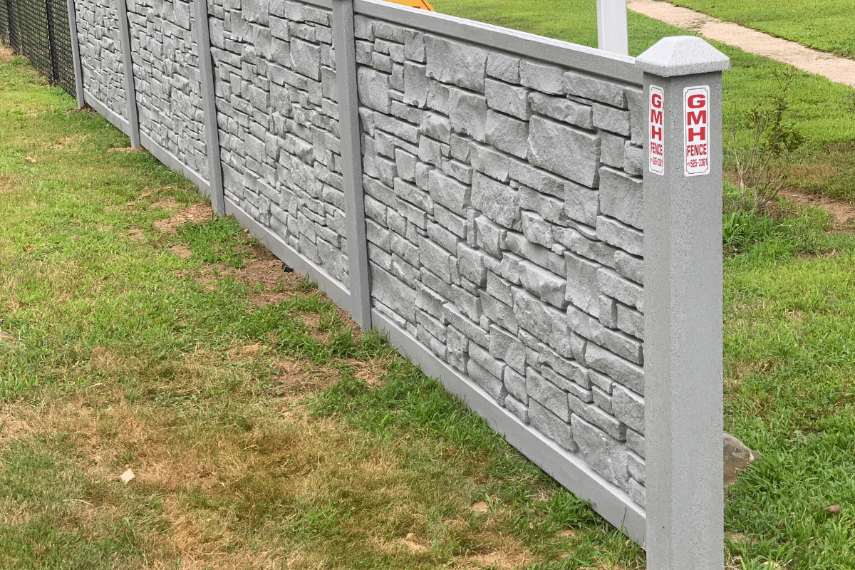 A gray vinyl, faux-stone fence runs along a grassy property in Meriden, CT. The fence posts has a red GMH FENCE label, and a sidewalk is visible in the background.