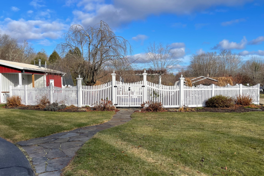 A curved stone pathway leads across a green lawn to a white picket fence and gate, with dormant shrubs and trees in a yard under a bright blue sky with scattered clouds.