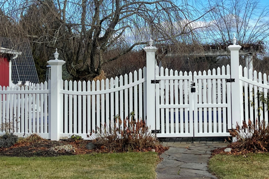 “White vinyl picket fence with tall posts and decorative caps, featuring a curved top section and double gate along a stone walkway in a landscaped yard”