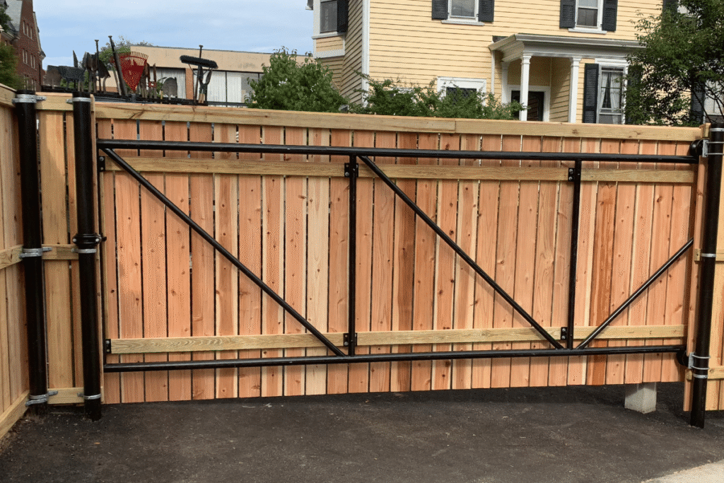 A wooden sliding gate with black metal supports stands closed at the entrance of a driveway, with a yellow house and greenery visible in the background.