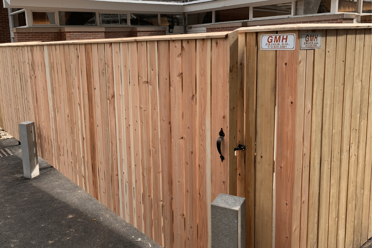 A tall wooden fence with a matching gate at UMass Amherst, featuring a black handle and two GMH contractor signs, runs alongside a paved road near a building with large windows and brick walls.