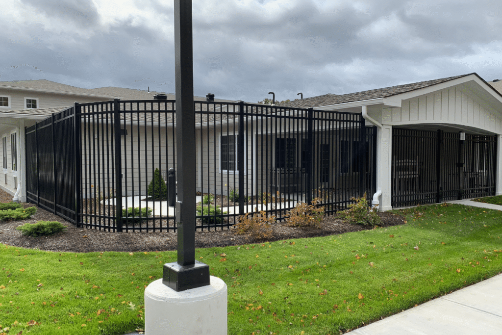 A black metal fence encloses a small courtyard area outside a tan building with white trim. There is some landscaping with bushes and grass, and a sidewalk runs alongside the fence under a cloudy sky.