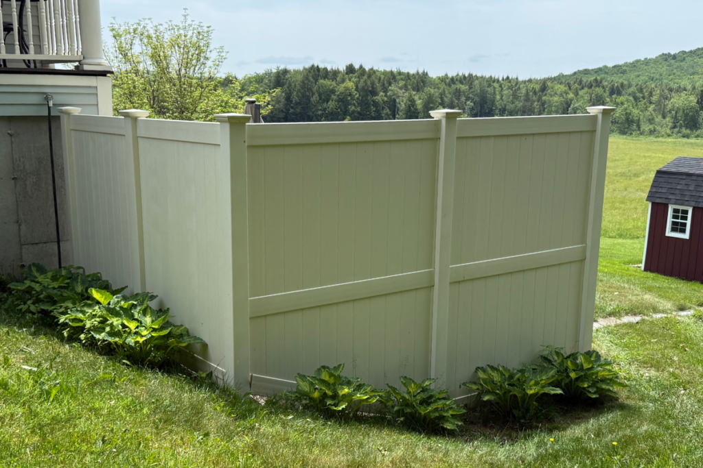 A beige privacy fence encloses a small area near a house. Green leafy plants grow along the base of the fence. In the background, there is a grassy field, trees, and a small red shed.