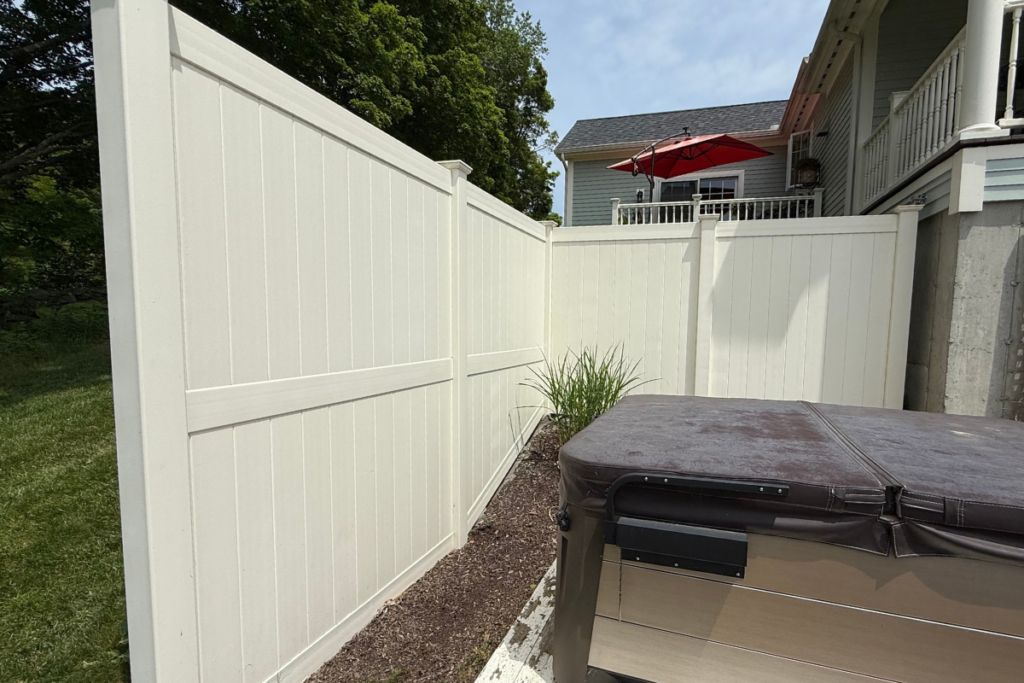 A tall white privacy fence encloses a corner of a backyard with a hot tub, some plants, and part of a house with a red patio umbrella visible on an upper deck.