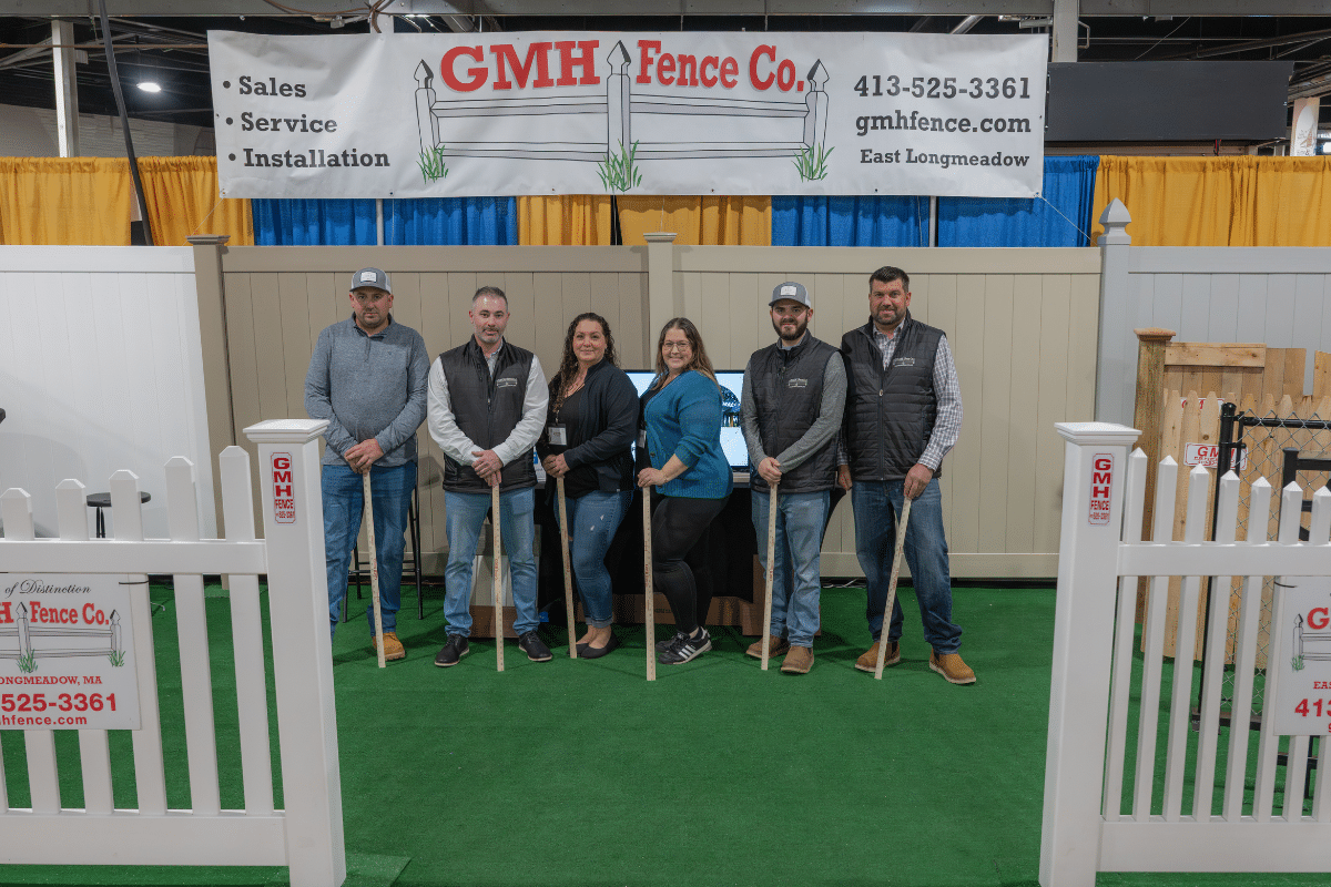 Six GMH team members stand in front of a fence display at The Original Western MA Home & Garden Show in West Springfield, MA. They are smiling and holding fence posts. A large banner with the company’s name and contact info hangs above them.