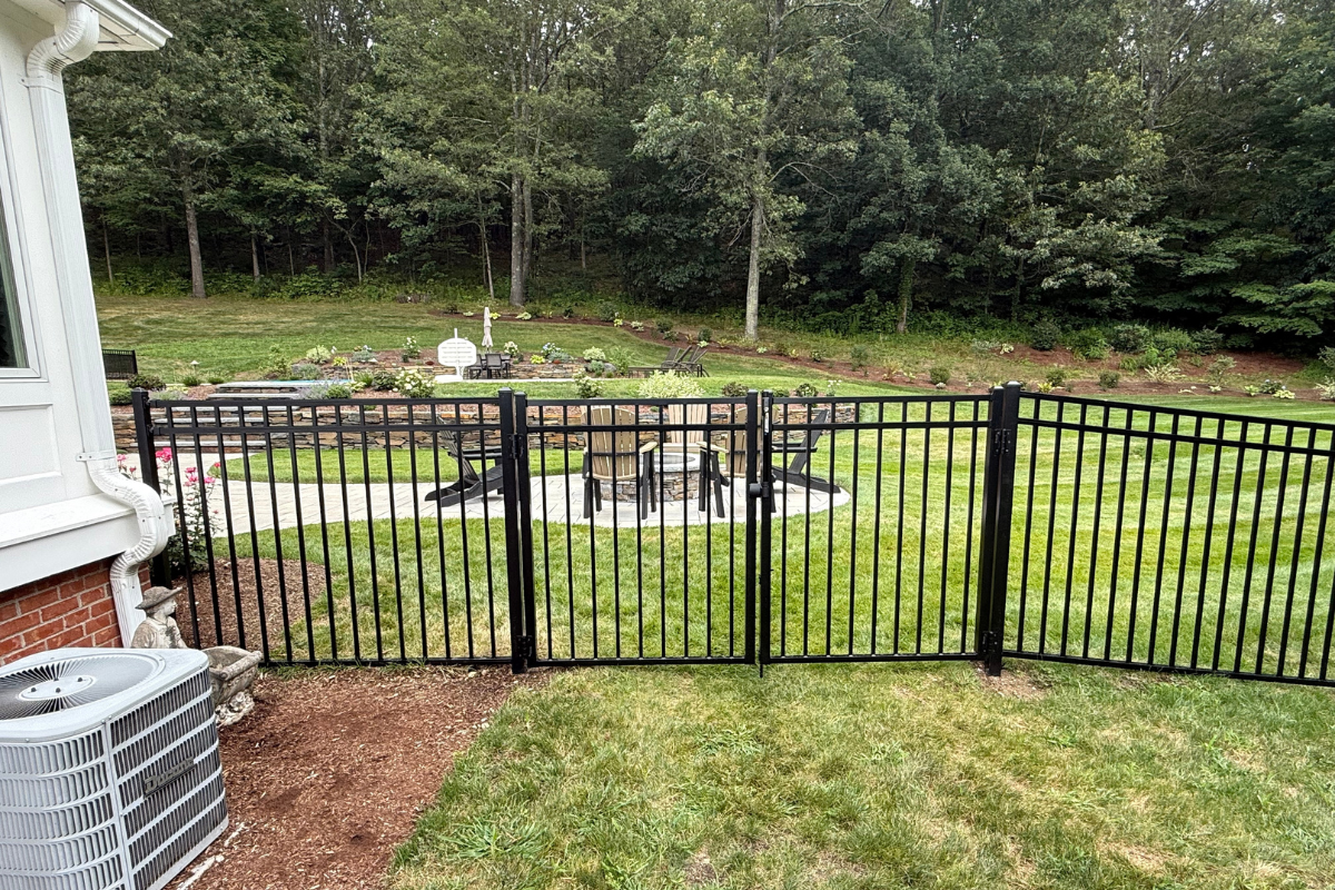 A black metal fence encloses a patio area with a table and chairs on a circular concrete pad, surrounded by green grass and trees in a spacious backyard.