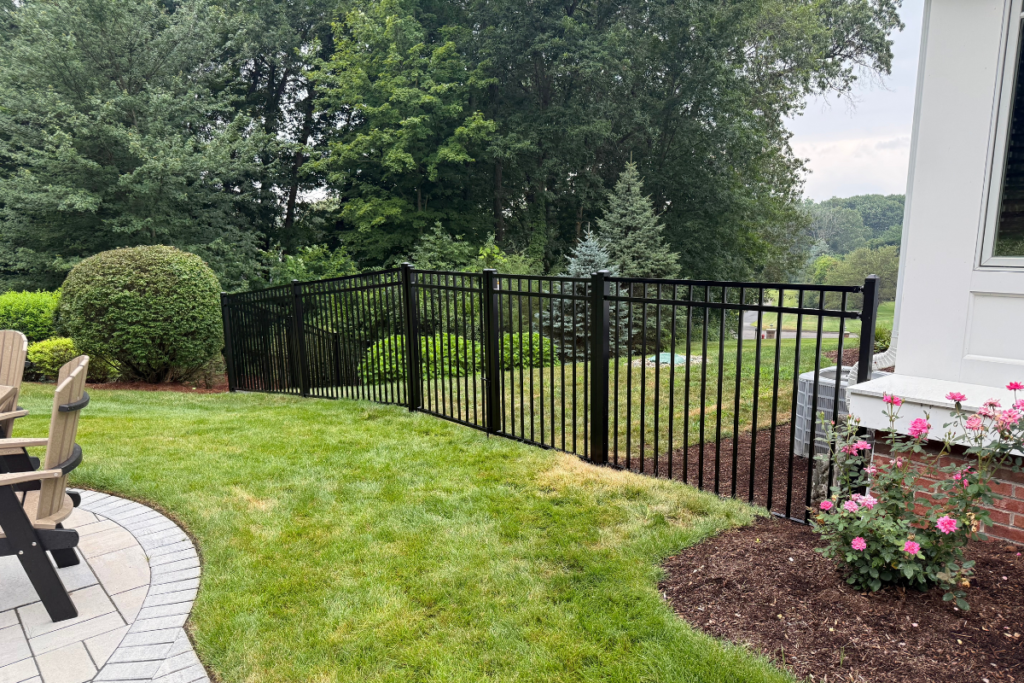 Black metal fence divides a green lawn with shrubs, trees, pink flowers, and a patio with chairs on the left; house exterior is partially visible on the right. Dense woods are in the background.