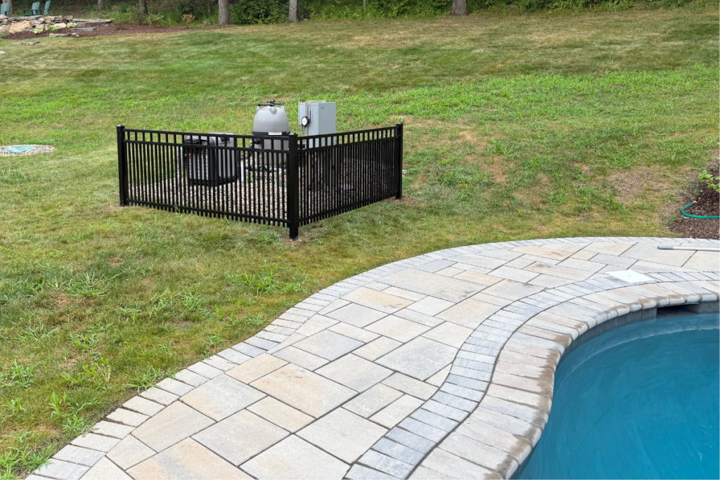 A fenced-in pool equipment area sits on grass near a paved patio and curved swimming pool, with a green lawn and trees in the background.