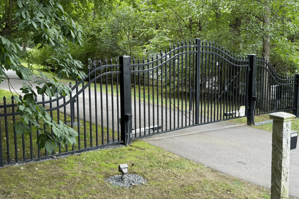A black metal gate with curved bars sits partially open across a paved driveway, surrounded by green trees and grass on a bright, clear day.