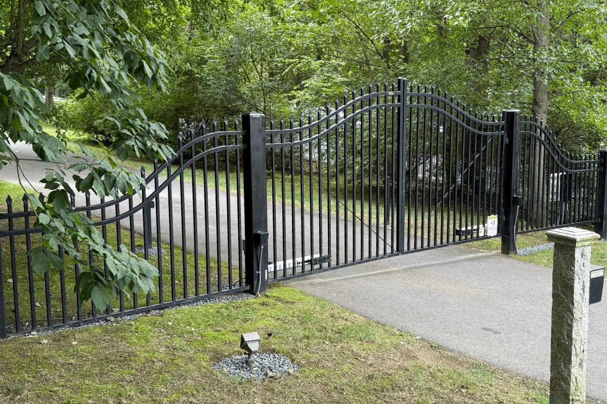 A black metal gate with curved bars sits partially open across a paved driveway, surrounded by green trees and grass on a bright, clear day.