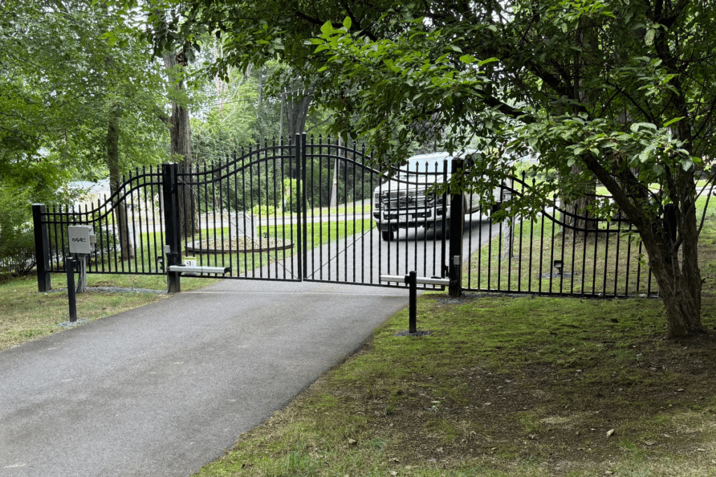 A black metal driveway gate stands open in a tree-lined yard, with a silver pickup truck parked just inside on the asphalt driveway. A mailbox and keypad are visible on the left side of the gate.
