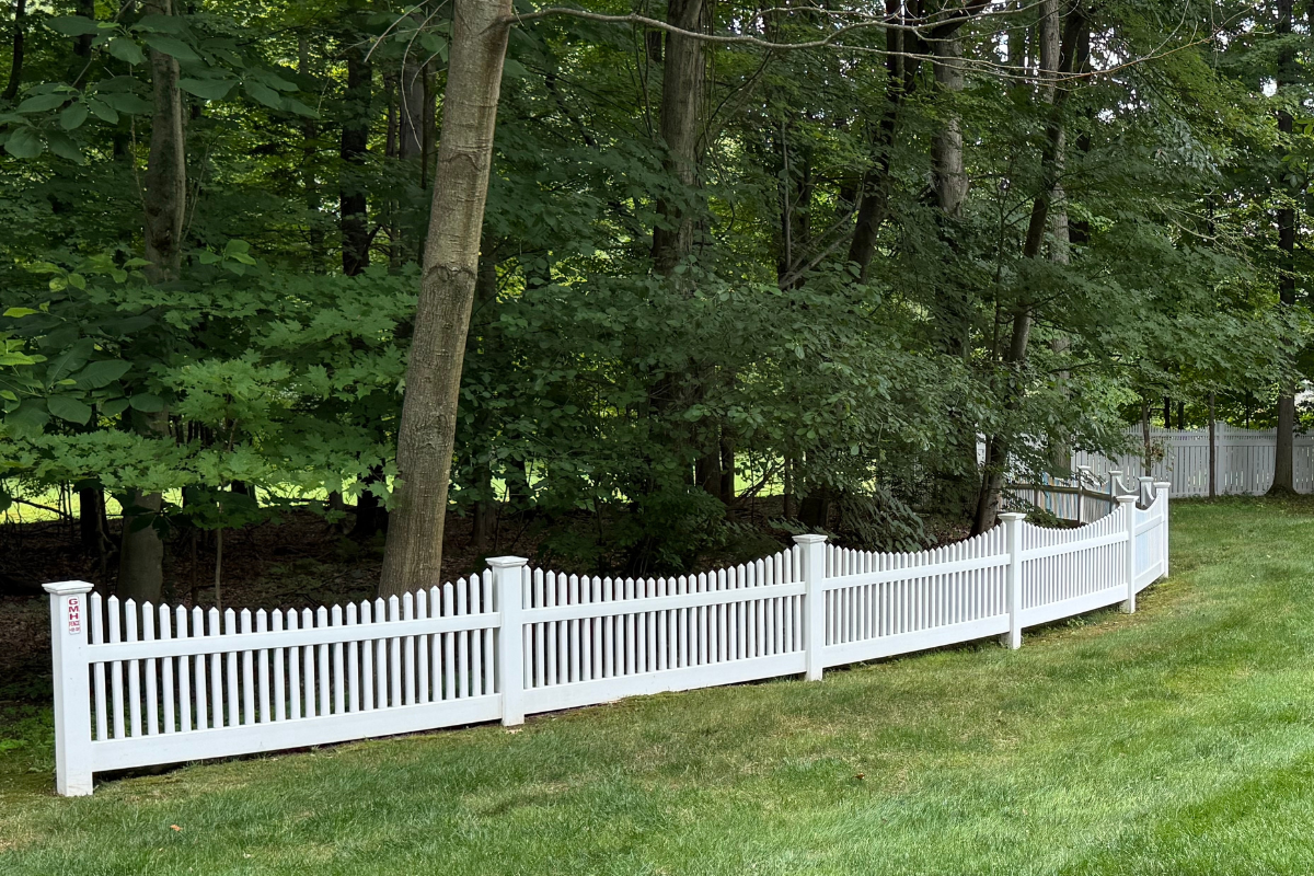 A white picket fence with sections that curve unevenly is set along the edge of a green lawn, bordering a wooded area with tall trees and dense foliage.