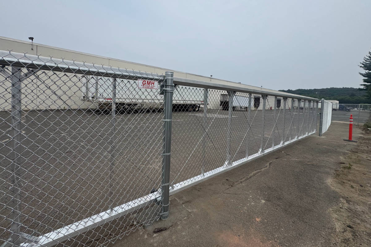 A large chain-link sliding gate blocks access to a fenced industrial lot at Cartamundi in East Longmeadow MA, with several white trucks parked beside a beige warehouse building in the background. The sky is overcast and some pavement is visible in the foreground.