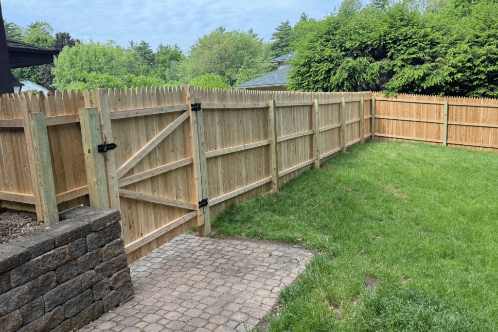 A wooden privacy fence with pointed tops and a gate stands next to a stone retaining wall, enclosing a grassy backyard with trees and shrubs in the background under a partly cloudy sky.