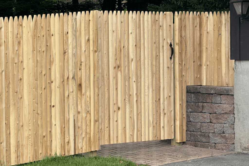 A tall wooden privacy fence with a gate and black handle stands next to a stone retaining wall and a paved walkway, with grass in the foreground.