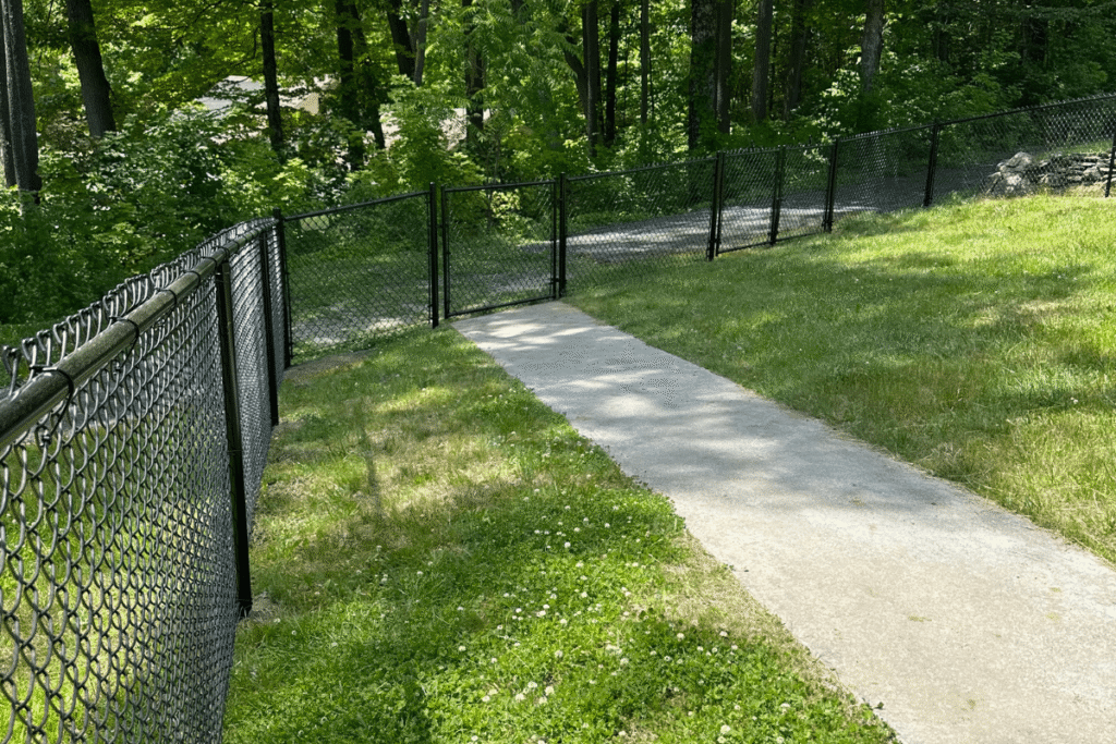 A narrow concrete path leads to a closed black chain-link gate, surrounded by green grass and trees in a wooded area on a sunny day.