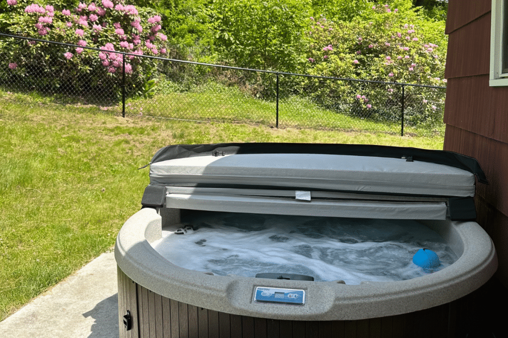 A bubbling outdoor hot tub with its cover partially open sits on a patio next to a house. A blue ball floats in the water. Green grass, blooming bushes, and a chain-link fence are in the background.