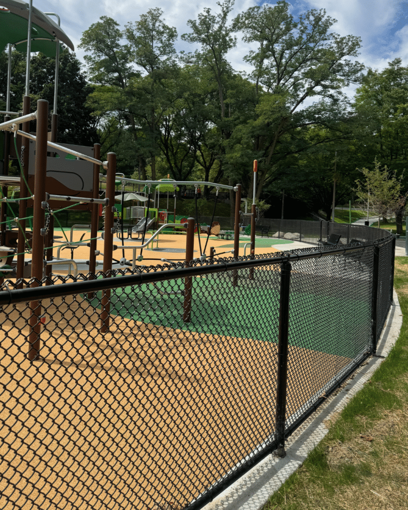 Outdoor playground with climbing structures and swings, surrounded by a black chain-link fence. The ground is covered with tan and green safety surfacing. Trees and grassy areas are visible in the background.