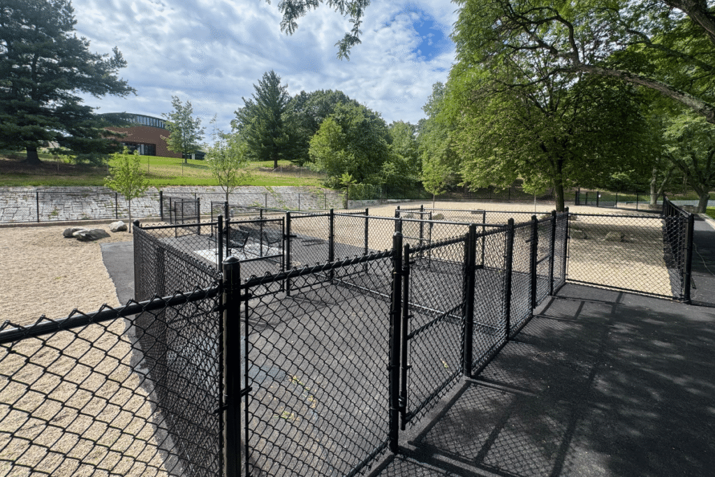 A fenced outdoor area with multiple gates, surrounded by trees and a sandy ground, under a partly cloudy sky.
