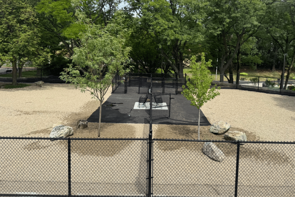 A fenced-in dog park with gravel ground, two young trees, large rocks, and benches in the center, surrounded by mature green trees.