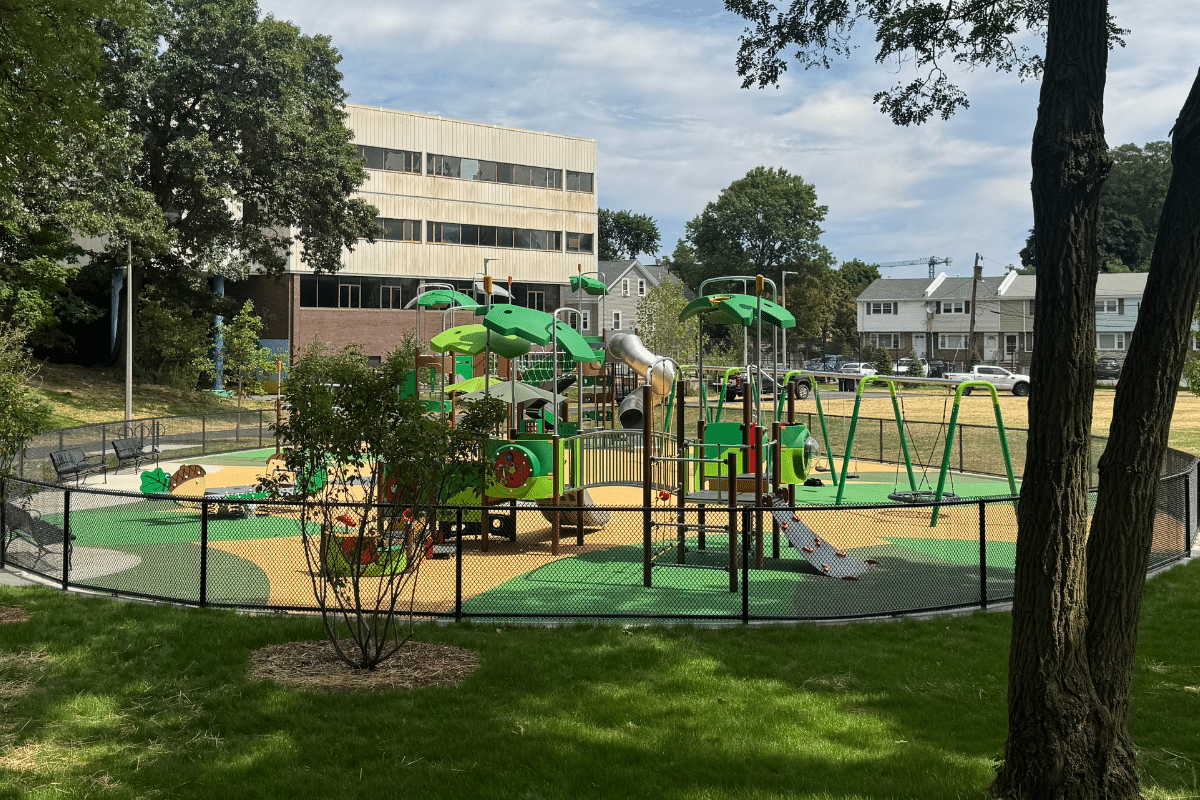 A fenced playground with green and yellow equipment, swings, a slide, and a climbing structure. Trees and grass surround the area, with an office building and houses visible in the background.